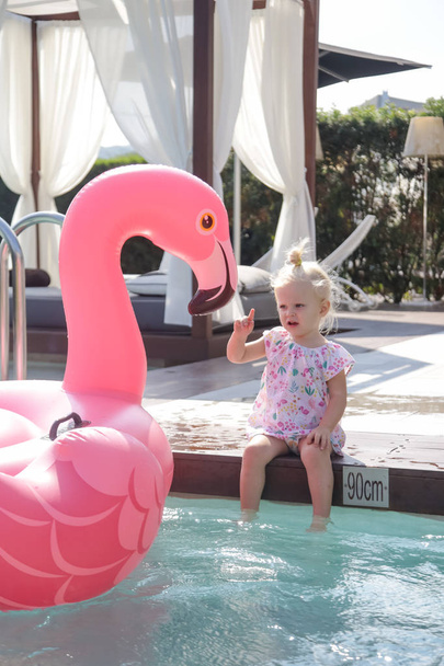 Αξιολάτρευτο κοριτσάκι που διασκεδάζει στην πισίνα και παίζει με μεγάλο ροζ φουσκωτό φλαμίνγκο - Φωτογραφία, εικόνα