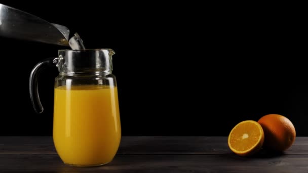 Wide shot of ice cubes falling from the scoop in decanter with fresh orange juice in slow motion on a wooden table decorated with oranges against black background - Materiaali, video