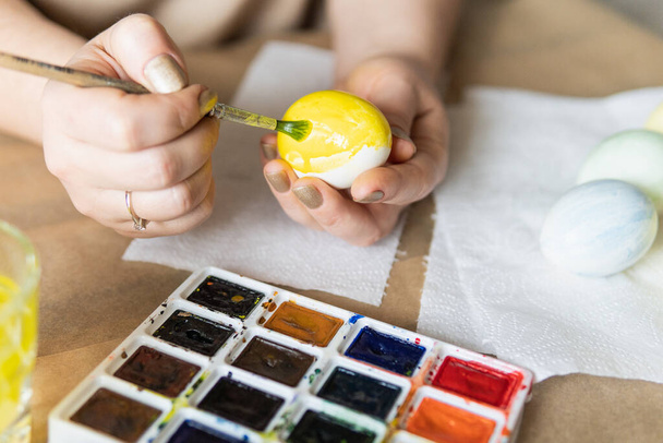 Young blonde woman painting eggs with watercolors preparing for Easter celebration. Closeup on hands - Фото, изображение