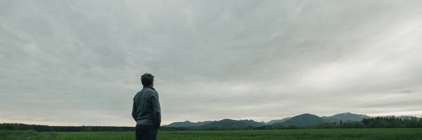 Ampia vista immagine di giovane in abbigliamento casual in piedi in bella natura sotto cielo grigio nuvoloso guardando in lontananza
. - Foto, immagini
