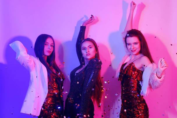 Three beautiful girls are dancing in neon. An incredibly fun party with girls in shiny dresses, they celebrate the holiday, smile, laugh, dance, sing. Night club dancing. Clapperboard, sweets. - Foto, afbeelding