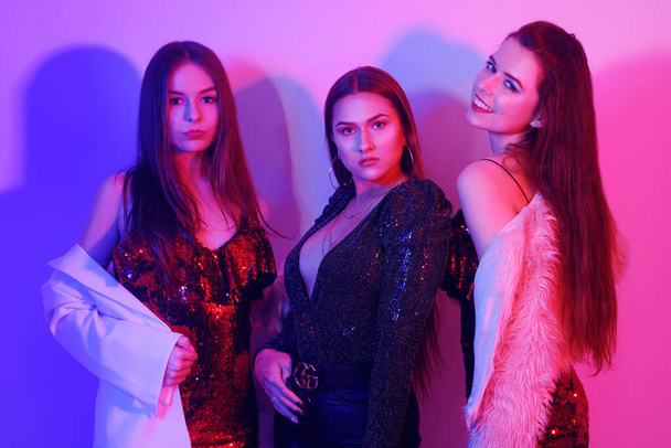 Three beautiful girls are dancing in neon. An incredibly fun party with girls in shiny dresses, they celebrate the holiday, smile, laugh, dance, sing. Night club dancing. - Valokuva, kuva