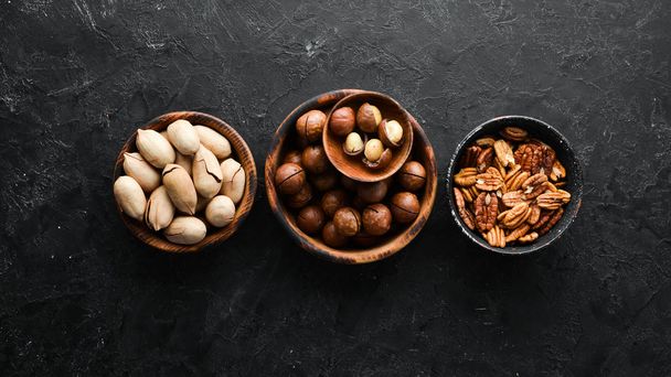 Exotic pecans and macadamia nuts in bowls on black stone background. Top view. Free space for your text. - Фото, изображение