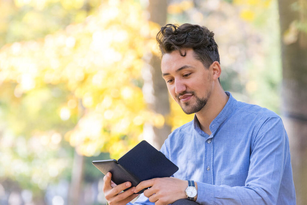 Un bel giovanotto che legge un ebook in un parco. Ritratto di un giovane uomo con giacca di jeans e camicia blu che legge un e-book all'esterno. Un ragazzo che legge un ebook in un parco
. - Foto, immagini