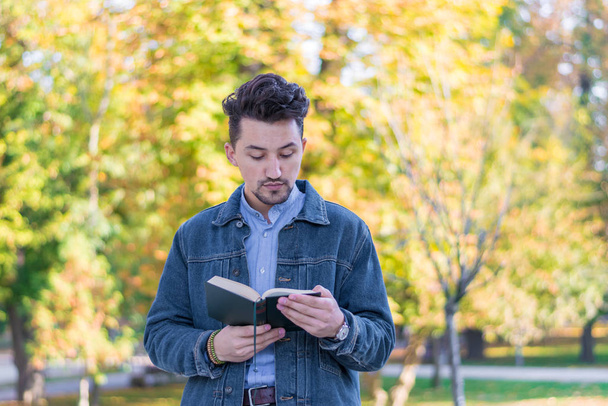 公園で本を読んでいるハンサムな若い男。デニムジャケットと青いシャツを着た若者が外で本を読んでいる姿が描かれています。公園で本を読んでいる男. - 写真・画像