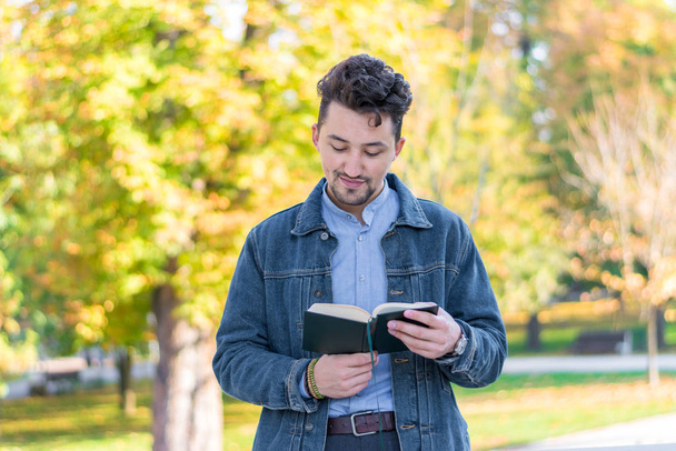 公園で本を読んでいるハンサムな若い男。デニムジャケットと青いシャツを着た若者が外で本を読んでいる姿が描かれています。公園で本を読んでいる男. - 写真・画像