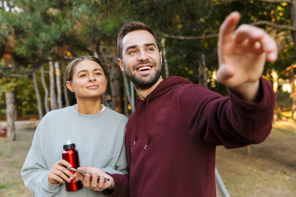 Picture of a happy positive sports woman and man using mobile phone outdoors in nature green park pointing aside holding bottle with water. - Foto, imagen