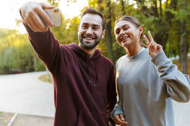 Photo of beautiful happy couple in sportswear taking selfie photo on cellphone and gesturing peace sign in green park - Fotó, kép