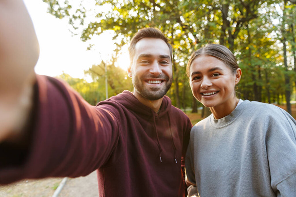 Photo of caucasian happy couple in sportswear taking selfie photo and smiling sign in green park - Zdjęcie, obraz