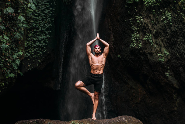 A man of athletic build does yoga. Healthy lifestyle. The concentration of the body. A man does yoga at a waterfall. A man does yoga in Bali. A man meditates in nature. Meditation at the waterfall - Fotografie, Obrázek