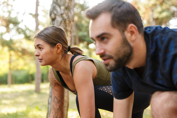 Foto de pareja joven y seria en ropa deportiva haciendo ejercicios mientras hace ejercicio en el parque verde durante el soleado día de verano.
 - Foto, Imagen