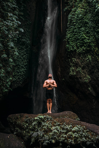 A man of athletic build does yoga. Healthy lifestyle. The concentration of the body. A man does yoga in nature. A man does yoga in Bali. A man meditates in nature. Meditation at the waterfall - Foto, immagini