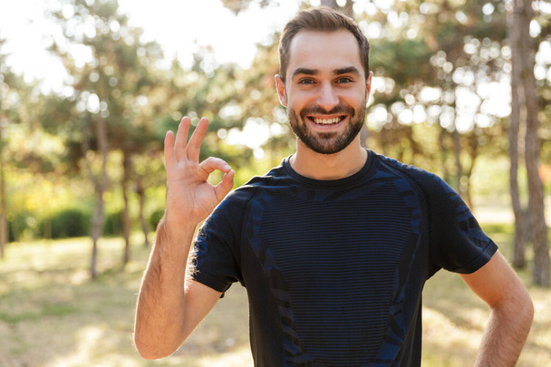 Image of a young strong pleased sports man posing outdoors in nature green park showing okay gesture. - Foto, Bild