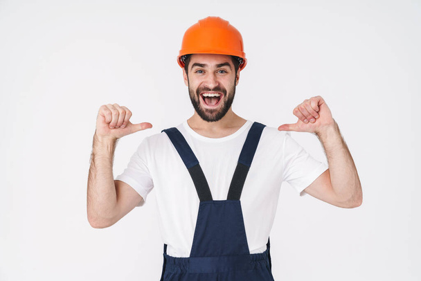 Photo of a cheery optimistic young man builder in helmet posing isolated over white wall background pointing to himself. - Fotografie, Obrázek