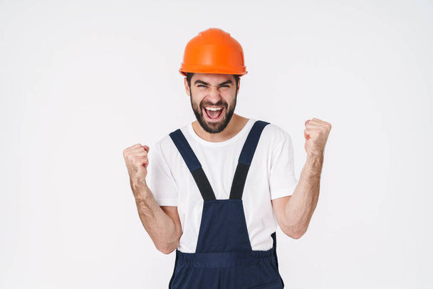 Photo of emotional young man builder in helmet posing isolated over white wall background make winner gesture. - Foto, Imagen