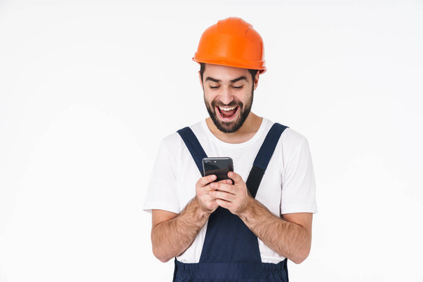 Image of a cheerful young man builder in helmet posing isolated over white wall background using mobile phone. - Valokuva, kuva