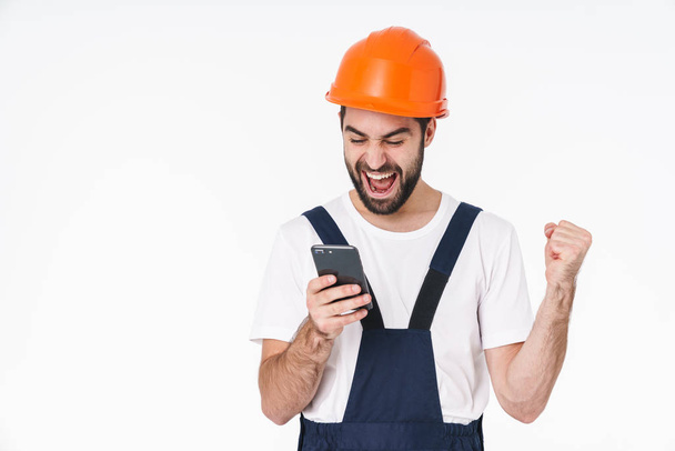 Photo of a happy positive young man builder in helmet posing isolated over white wall background using mobile phone. - Photo, Image