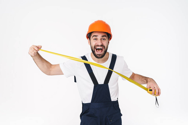 Fotografie pozitivního šťastný šokovaný mladý muž stavitel v helmě pózování izolované přes bílou zeď pozadí držení centimetr. - Fotografie, Obrázek
