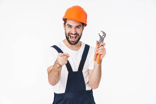 Image of happy young man builder in helmet posing isolated over white wall background holding plumbing wrench pointing to you. - Valokuva, kuva