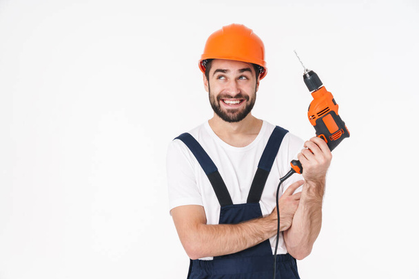 Photo of cheerful young man builder in helmet posing isolated over white wall background holding drill. - Valokuva, kuva