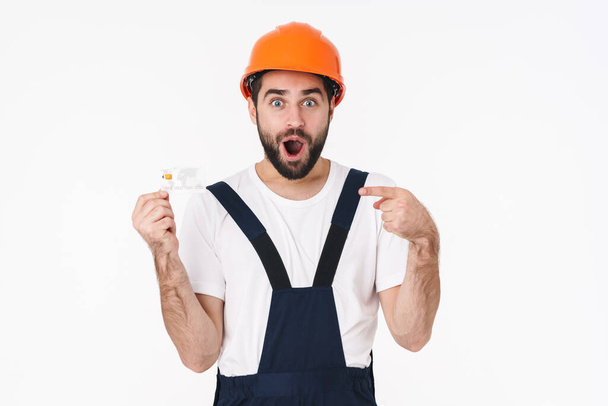 Imagen del conmocionado joven constructor emocional en casco aislado sobre fondo de pared blanca sosteniendo tarjeta de crédito apuntando
. - Foto, Imagen
