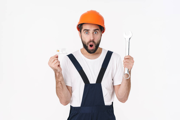 Image of shocked young man builder in helmet isolated over white wall background holding credit card and wrench. - Фото, изображение