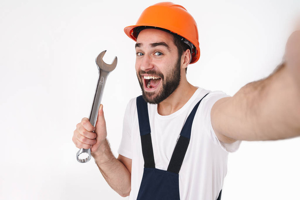 Image of happy positive young man builder in helmet isolated over white wall background take selfie by camera holding wrench. - Foto, Bild