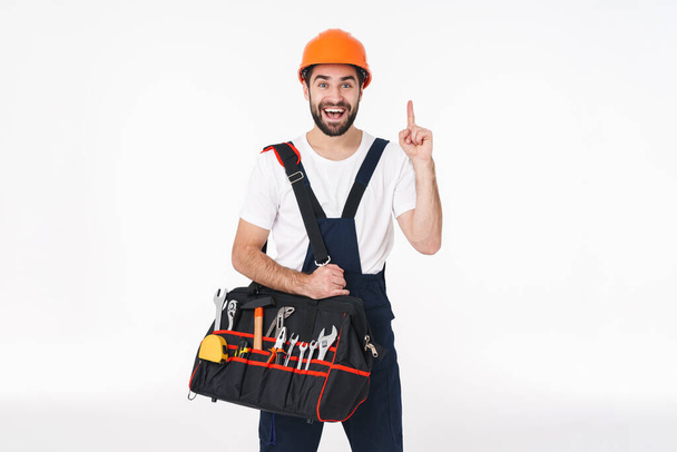 Image of excited surprised young man builder in helmet isolated over white wall background holding bag with equipment instruments have an idea. - Photo, Image