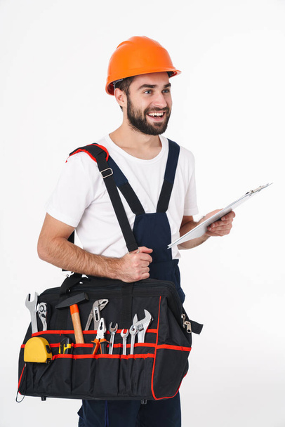 Photo of happy young man builder in helmet isolated over white wall background holding bag with equipment instruments holding clipboard. - Photo, image