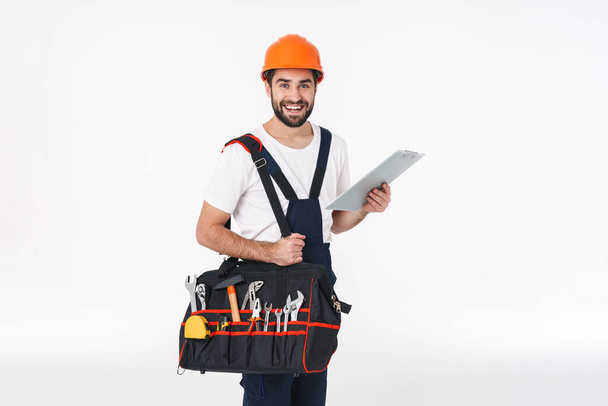 Image of smiling positive young man builder in helmet isolated over white wall background holding bag with equipment instruments holding clipboard. - Φωτογραφία, εικόνα