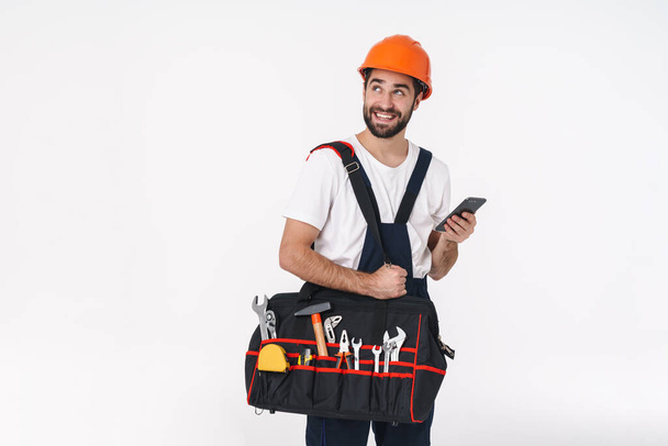 Image of smiling young man builder in helmet isolated over white wall background holding bag with equipment instruments using mobile phone. - Фото, изображение