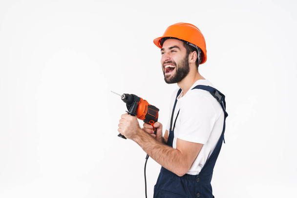 Image of emotional young man builder in helmet isolated over white wall background holding drill. - Φωτογραφία, εικόνα