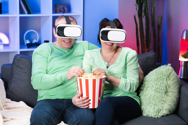 Old Caucasian Spouses Sitting Together at Sofa Eating Popcorn and Watching Movie in VR Goggles. Family couple is sitting at the sofa with popcorn and watching TV using VR glasses. - Fotografie, Obrázek