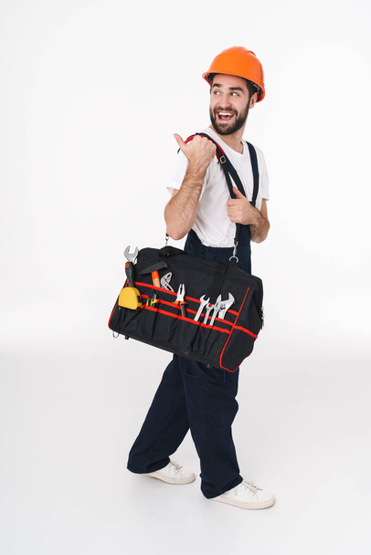 Picture of positive emotional young man builder in helmet isolated over white wall background holding bag with equipment instruments pointing aside. - Φωτογραφία, εικόνα