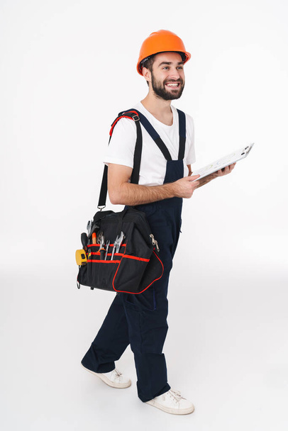 Picture of cheerful smiling young man builder in helmet isolated over white wall background holding bag with equipment instruments and clipboard. - Foto, afbeelding