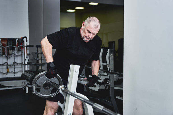 Adult gray-haired man trains on fitness equipment in the gym, pumps legs and arms muscles, loses weight. Concept of healthy lifestyle in old age - Foto, immagini
