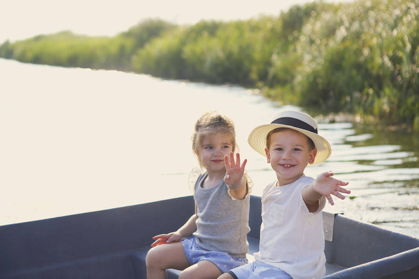 Cheerful boy and girl smiling and waving hands while sitting in boat in calm river in countryside - Photo, Image