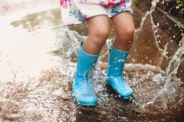 Játékos izgatott óvodás gyermek alkalmi ruhák és gumicsizma kék esernyő nevet, és ugrál a tócsa mosolygó kamera közben játszik a parkban - Fotó, kép