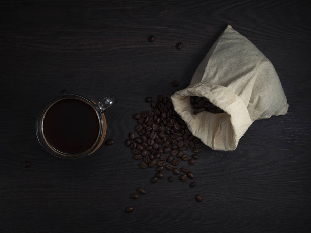 glass mug with fresh black natural coffee, a bag with coffee grains and sprinkled grains of roasted coffee on a wooden table. - Фото, зображення