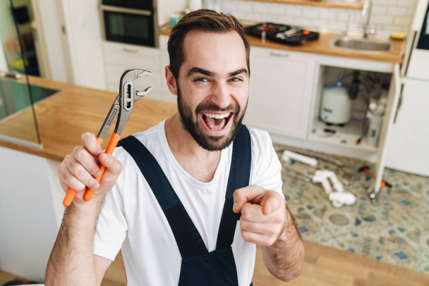 Image of young positive happy man plumber work in uniform indoors holding pipe wrench pointing to you. - Zdjęcie, obraz