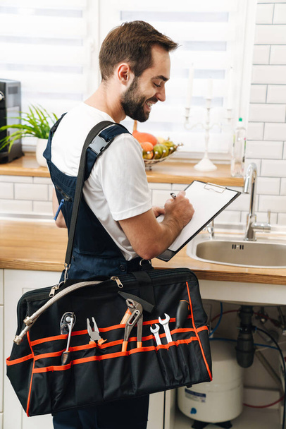 Image of young man plumber work in uniform indoors holding bag with equipment and clipboard. - Photo, image