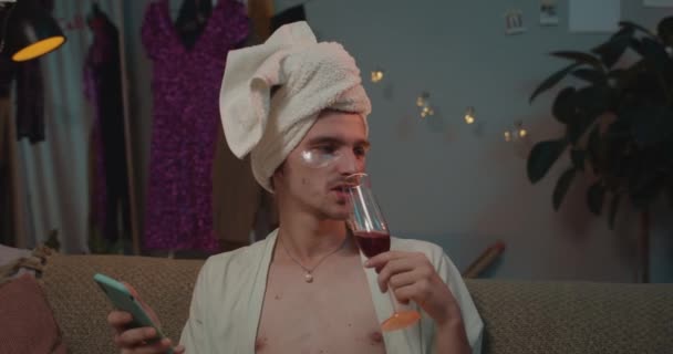 Close up of trans man in home wear using smartphone and listening to music. Relaxed transgender man in undereye patches sitting on couch at home enjoing free time and savoring wine. Indoor. - Séquence, vidéo