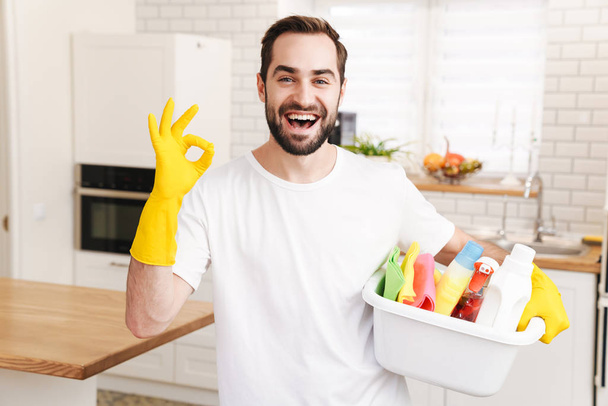 Image of young optimistic positive man househusband indoors showing okay gesture. - Foto, imagen