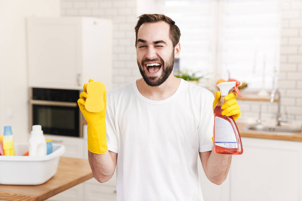 Image of young cheerful smiling positive man househusband indoors winking holding sponge and cleansing spray. - Foto, imagen