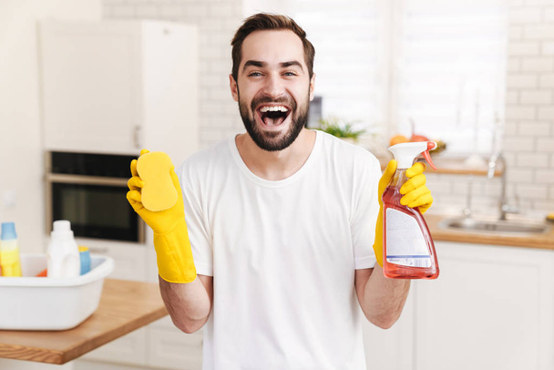 Image of young optimistic positive man househusband indoors holding sponge and cleansing spray. - Фото, изображение