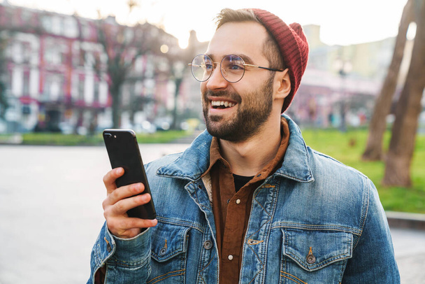 Close up of a handsome happy young stylish bearded man walking outdoors in the street, using mobile phone - 写真・画像