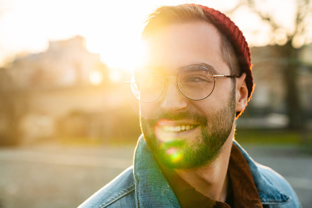 Close up of a handsome happy young stylish bearded man walking outdoors in the street, looking away - 写真・画像