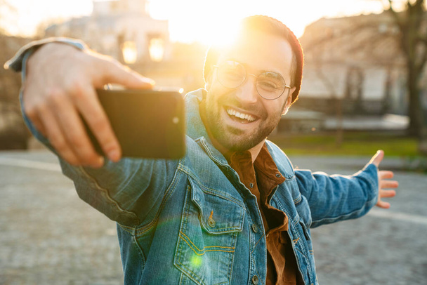 Close up of a handsome happy young stylish bearded man walking outdoors in the street, taking a selfie - Foto, Bild