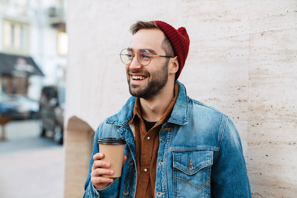 Close up of a handsome happy young stylish bearded man walking outdoors in the street, holding takeaway coffee cup - Valokuva, kuva