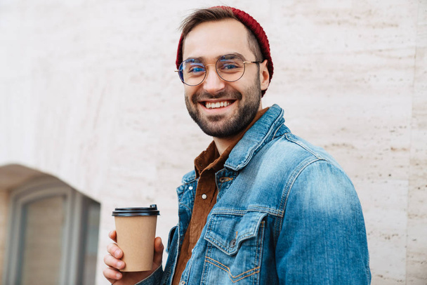 Close up of a handsome happy young stylish bearded man walking outdoors in the street, holding takeaway coffee cup - Φωτογραφία, εικόνα